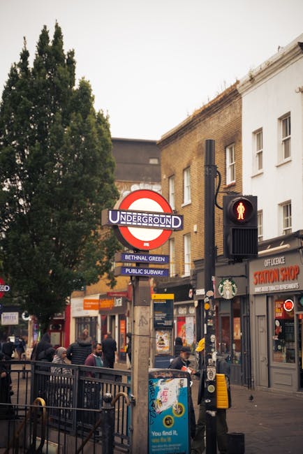 A busy outdoor scene at Bethnal Green with a prominent London Underground station sign featuring the classic red circle and blue bar, indicating the station name. The area is lively with pedestrians w